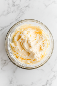 This image shows softened cream cheese being added to a lemon mixture in a clear glass bowl, ready to be blended into a smooth filling on a white marble countertop.