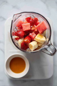 adding_frozen_fruits_and_honey_to_glass_blender_on_white_marble