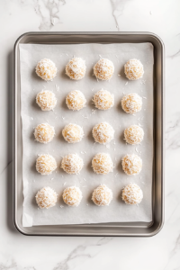 This image shows coconut-coated Apple Coconut Bites arranged in neat rows on a parchment-lined baking sheet placed over a white marble cooktop.