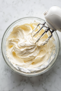 beating-cream-cheese-in-clear-glass-bowl-with-hand-mixer-on-white-marble