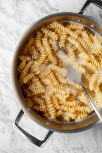 This image shows rotini pasta cooking in boiling salted water inside a large stainless steel stockpot placed on a clean white marble countertop.