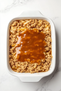 This image shows thick golden caramel sauce being poured and smoothed over the layered cake mixture inside a white ceramic dish placed on a white marble countertop.