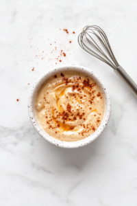 This image shows a small white ceramic bowl of creamy chili-lime dressing on a white marble countertop, with visible spices and a whisk beside it.