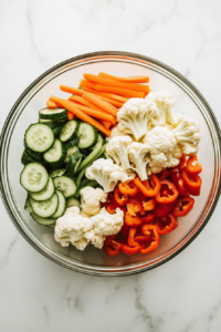 This image shows a glass bowl filled with colorful chopped vegetables—cucumbers, cauliflower, carrots, bell peppers, and green beans—ready for pickling.