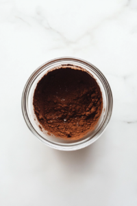 This image shows cocoa powder mixed with warm water into a paste in a small glass bowl on a white marble countertop.