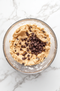 This image shows the cookie dough mixed with flour and chocolate chips in a silver stand mixer bowl on a white marble cooktop.