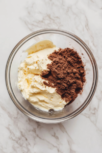 This image shows cream cheese and sugar being combined with cocoa paste in a transparent glass bowl on a white marble surface.