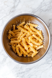 This image shows freshly boiled rotini pasta sitting in a stainless steel colander over a white marble sink, drained and ready to cool.