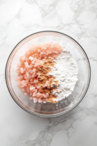 This image shows a large glass bowl on a white marble countertop, filled with chopped rhubarb, sugar, flour, water, cinnamon, and allspice mixed together.