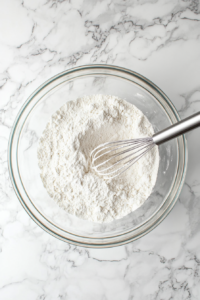 glass-bowl-This image shows a glass bowl filled with flour, cornstarch, baking soda, and salt on a white marble cooktop, ready to be whisked together.with-dry-ingredients-for-mini-cookie-recipe