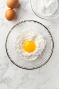 This image shows a clear glass bowl filled with unmixed milk cookie ingredients including 1 egg, sugar, shortening, baking soda, cream of tartar, nutmeg, salt, and sweetened condensed milk, resting on a clean white marble countertop.