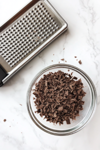 This image shows a glass bowl of finely grated dark chocolate with a microplane grater and chocolate bar on a white marble countertop.