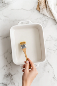 This image shows a greased white ceramic baking dish on a clean white marble countertop, with a bottle of oil and red-bristled silicone brush beside it.