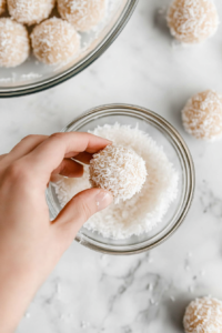 This image shows dough balls being coated in shredded coconut inside a white ceramic bowl placed on a white marble cooktop.