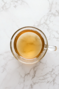 This image shows the same transparent glass mug placed on a white marble countertop, containing a warm mixture of water and melted honey after microwaving.