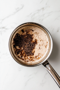 This image shows a stainless steel saucepan on a white marble cooktop containing whole milk and brewed coffee being gently heated.melting-chocolate-in-coffee-and-milk-mixture-on-white-marble