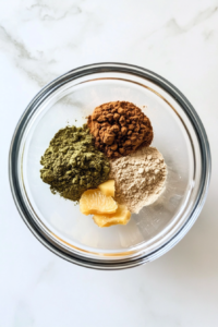 This image shows a top-down view of a clear glass bowl containing ceremonial-grade matcha powder, powdered kelp, ground ginger, and dried yuzu peel arranged neatly before mixing.