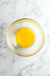 This image shows a glass mixing bowl with fully melted golden butter sitting on a clean white marble countertop.
