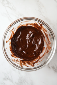 This image shows melted chocolate in a transparent glass bowl on a white marble countertop with a clean and simple background.