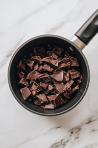 This image shows chopped dark chocolate melting in a black saucepan over white marble, in the process of making ganache.