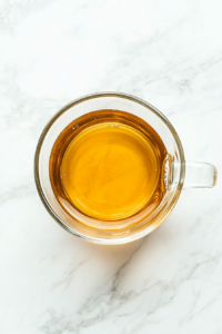 This image shows a transparent glass microwave-safe mug filled with 1 cup of water, placed on a clean white marble countertop. The scene is simple, focused solely on the mug and water.