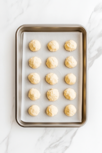 This image shows round milk cookie dough pieces arranged neatly on a parchment-lined aluminum baking sheet over a white marble countertop, ready for baking.