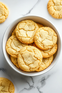 This image shows golden, sugar-dusted milk cookies stacked in a glossy white ceramic bowl placed on a clean white marble countertop with no background clutter.