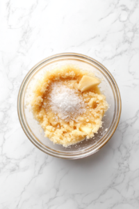 This image shows apple pie filling mixed with lemon juice, vanilla extract, and kosher salt in a clear glass bowl over a white marble countertop.
