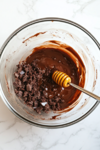This image shows a clear glass bowl on a white marble surface, containing crushed chocolate cake, melted dark chocolate, and honey being combined into a rich dough.