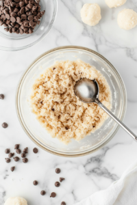 This image shows a glass bowl on a white marble cooktop containing sweetened condensed milk, graham cracker crumbs, shredded coconut, chocolate chips, and vanilla extract ready to be mixed.
