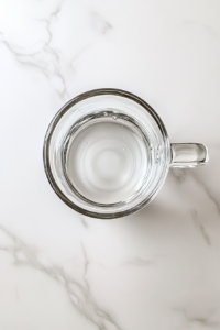 This image shows a transparent glass mug on a clean white marble surface, now containing water and 1 tablespoon of golden honey, clearly visible at the bottom of the mug.