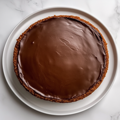 This image shows a finished no bake chocolate cheesecake, perfectly smooth and glossy, set on a clean white marble countertop with no other objects in the background.