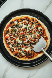 This image shows an offset spatula placed beside a pizza crust already layered with bean mixture on a black tray over white marble.