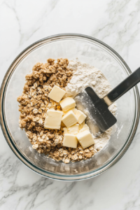 This image shows a glass bowl with a pastry cutter inside, blending butter into the oat, brown sugar, and flour mixture to form a crumbly topping.