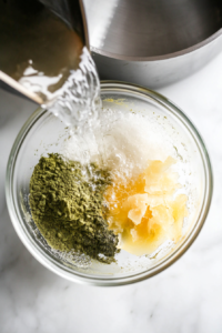 This image shows a top-down view of hot water just poured into a glass bowl containing matcha powder, kelp, ginger, and yuzu peel on a white marble cooktop.