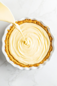 This image shows smooth lemon pie filling being poured into a graham cracker crust in a white ceramic pie dish, resting on a white marble countertop.