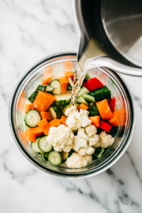 This image shows warm pickling brine being poured into a glass jar filled with assorted vegetables, as part of the mixed pickle preparation.