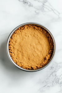 This image shows a graham cracker crust pressed into a silver springform pan, resting on a clean white marble countertop.