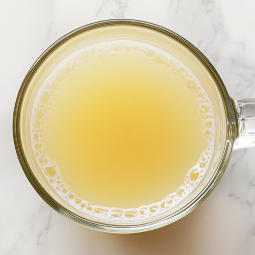This image shows a clear glass filled with lemon honey drink, placed on a clean white marble countertop. The pale yellow drink gleams under natural light, showcasing its simplicity and refreshment. A white background ensures nothing distracts from the featured beverage.