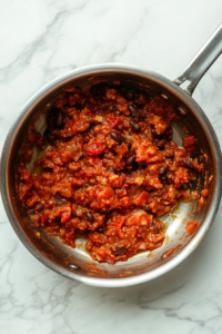 This image shows a silver saucepan containing refried black beans being heated gently on a clean white marble cooktop.