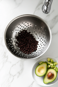 This image shows black beans being rinsed in a small stainless steel colander and freshly diced avocado on a white plate, all set on a white marble countertop.