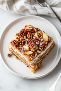This image shows a square slice of the caramel cream cake garnished with flaky salt and chopped pecans, served on a round white ceramic plate over a white marble countertop.