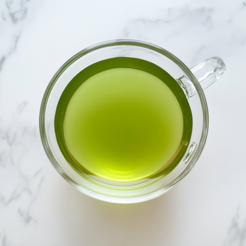 This image shows a top-down view of finished Japanese Mounjaro tea in a clear glass bowl with raw honey blended in, set on a clean white marble surface.