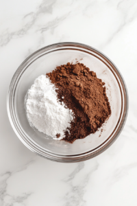 This image shows dry ingredients for mocha frosting—confectioners’ sugar, cocoa powder, and espresso powder—sifted into a glass bowl.