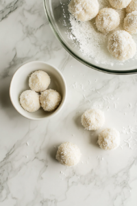 This image shows the sticky dough in a glass bowl with a few rolled Apple Coconut Bites placed beside it on a white marble cooktop.