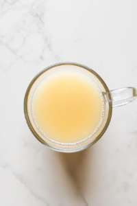 This image shows a clear glass mug placed on a clean white marble countertop, now containing warm honey water with 1 tablespoon of lemon juice added, giving it a slightly cloudy pale yellow color.