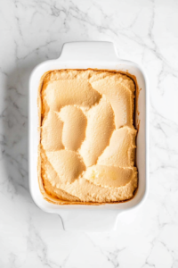 top-down-adding-creamy-hummus-to-broth-mixture-in-white-baking-dish-on-marble