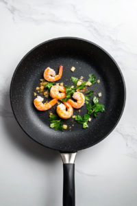 This image shows pre-cooked shrimp being lightly sautéed with garlic and parsley in a black skillet over a clean marble countertop.