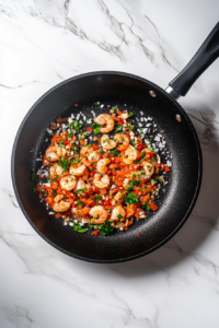 This image shows pasta and shrimp being tossed together in a skillet until coated in a light sauce, all placed over a clean marble cooktop.
