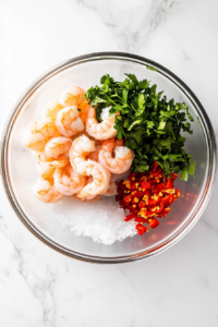 This image shows raw shrimp, minced garlic, parsley, and seasonings in a glass bowl with olive oil, set over a white marble cooktop.